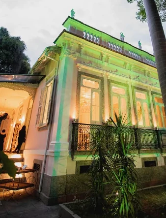Fachada do casarão iluminada em verde ao entardecer, com varanda de ferro trabalhado e palmeiras